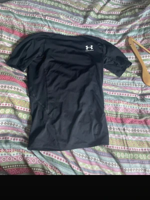 Svart kompressionströja Under Armour - Säljer en svart kortärmad kompressionströja från Under Armour med HeatGear-teknologi. Tröjan har en tight passform och är tillverkad i ett stretchigt funktionsmaterial som transporterar bort fukt. Perfekt för träning eller sport.