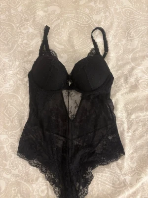 Svart spetsbody med vadderade kupor - Snygg svart body i transparent spets med blommigt mönster och vadderade kupor o push up. Justerbara axelband och djup urringning framtill. Bara använt en gång alltså inga defekter eller så💕