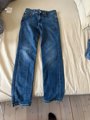 Blå Jack and Jones jeans model glen - Säljer ett par blå jeans med rak passform och klassisk femficksdesign. Jeansen har normal midja och är tillverkade i denim med lätt tvättad look. Perfekta för en avslappnad stil och passar till det mesta.