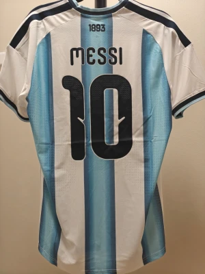 Argentina spelar version Messi matchtröja Adidas - Säljer Argentina fotbollströja från Adidas med Messi och nummer 10 på ryggen i spelar version. Tröjan har klassiska blå och vita ränder, guldiga detaljer och FIFA-märke på bröstet. Perfekt för dig som älskar fotboll och vill stötta landslaget.