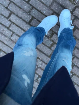Blå slitna jeans från replay - Säljer ett par blå jeans från replay med slitningar och ljusa tvättade partier. Jeansen har klassisk femficksmodell, låg midja och raka ben/ lite bootcut.