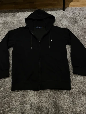 Svart hoodie från Polo Ralph Lauren - Svart hoodie med dragkedja från Polo Ralph Lauren. Klassisk design med huva och dragsnören, samt den ikoniska vita logotypen broderad på bröstet. Mjuk bomullsblandning och ribbade muddar för en skön passform.