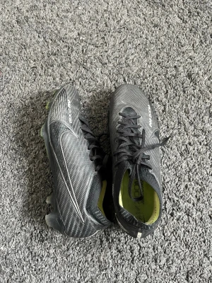 Nike Mercurial Vapor 14 fotbollsskor - Säljer ett par svarta Nike Mercurial Vapor 14 fotbollsskor med vita detaljer och snörning. Skorna har en modern, aerodynamisk design och är tillverkade i syntetmaterial med textur för bättre bollkontroll. Yttersulan har fasta dobbar för spel på gräs.