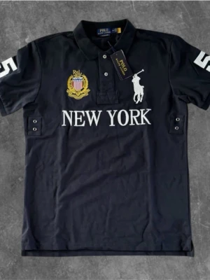 Polo Ralph Lauren Marinblå  pikétröja  - Mörkblå pikétröja från Polo Ralph Lauren 🧼med vit broderad logga och texten 'NEW YORK' framtill. Tröjan har klassisk krage, knappar vid halsen och detaljer på ärmarna. Perfekt för en sportig och stilren look.🐧 Priset är inte satt på sten 