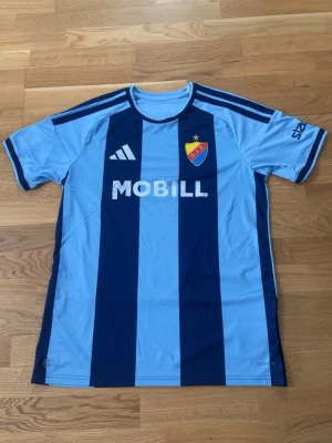 Djurgården matchtröja Adidas blå - Snygg Djurgården matchtröja från Adidas i ljusblått och mörkblått med vertikala ränder. Tröjan har klubbmärke på bröstet, sponsorloggor och korta ärmar. Perfekt för fotbollsträning eller att bära på läktaren. Tillverkad i lätt och ventilerande material.