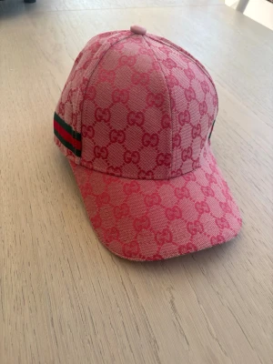 Rosa G keps med GG-mönster - Säljer en rosa keps från GG med klassiskt GG-monogram i hela tyget. Kepsen har en böjd skärm och justerbart spänne bak i rosa läder. På sidan finns GGS ikoniska gröna och röda ränder. Perfekt för dig som vill sticka ut med en lyxig accessoar.