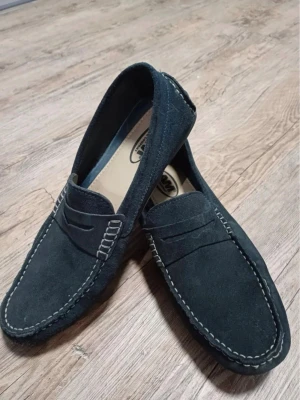 Mörkblå mockaloafers från AM Shoe - Snygga mörkblå loafers i mocka från AM Shoe Company. Skorna har klassisk design med dekorativa sömmar i vitt och en stilren pennyloafers-look. Perfekta för dig som gillar en clean och tidlös stil.