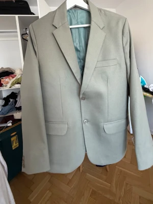 Ljusgrå klassisk kavaj - Ljusgrå kavaj med två knappar framtill, klassiska slag och tre fickor. Kavajen har rak passform och är helfodrad med ljusgrönt foder. Perfekt för dig som vill ha en stilren och enkel look. Materialet känns som en blandning av polyester och bomull.