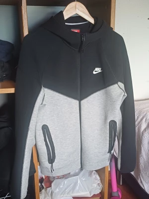 Nike Tech Fleece hoodie svart/grå - Nike Tech Fleece hoodie i svart och ljusgrått med dragkedja framtill, två fickor med dragkedja och klassisk Nike-logga på bröstet. Hoodien har en modern passform och är tillverkad i mjukt fleecematerial. Perfekt för en sportig och avslappnad stil.