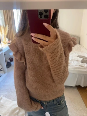 Beige stickad tröja med volang - Mysig beige stickad tröja med volangdetaljer längs axlarna. Tröjan har rund halsringning och långa ärmar med ribbade muddar. Perfekt för dig som gillar en söt och trendig look till vardags.
