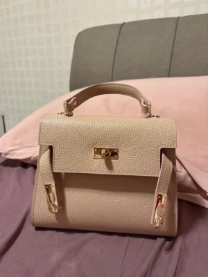 Beige handväska i äkta läder - Snygg beige handväska i äkta läder med guldiga metalldetaljer och klassisk topphandtag. Stilren och strukturerad design med lock och spänne framtill. Perfekt accessoar för en trendig look.