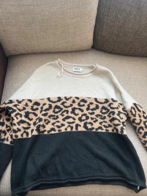 Leopardmönstrad stickad tröja ONLY - Stickad tröja från ONLY med beige upptill, leopardmönstrat parti i mitten och svart nedtill. Tröjan har rund halsringning och långa ärmar. Perfekt för dig som gillar djurmönster och vill sticka ut lite extra.