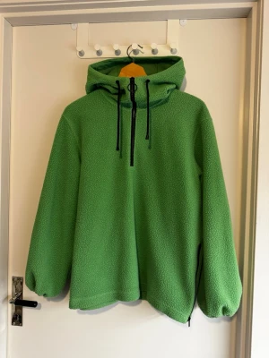 Grön fleecetröja med huva från Arket - Mysig grön teddyjacka/fleecetröja med huva och svart dragkedja framtill. Jackan har dragsko i huvan och en avslappnad passform. Tillverkad i fluffigt teddy-material som håller dig varm och bekväm. Perfekt för dig som gillar en chill och trendig stil. Ganska stor i storlek. 