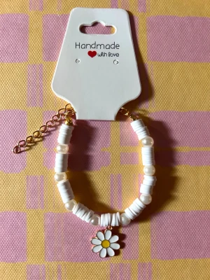 Vitt pärlarmband med blomberlock - Sött armband med vita platta pärlor och runda pärlor, dekorerat med en emaljerad blomberlock i vitt och gult. Armbandet har guldfärgad kedjeförlängning och är handgjort. Perfekt för dig som gillar gulliga accessoarer.