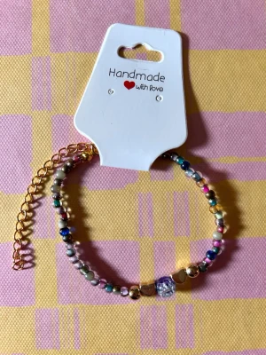 Färgglatt pärlarmband med gulddetaljer - Ett handgjort armband med små färgglada pärlor i olika nyanser och guldfärgade detaljer. Armbandet har en justerbar kedja i guldton och en unik glittrig pärla i mitten som blickfång. Perfekt accessoar för att lyfta din stil.