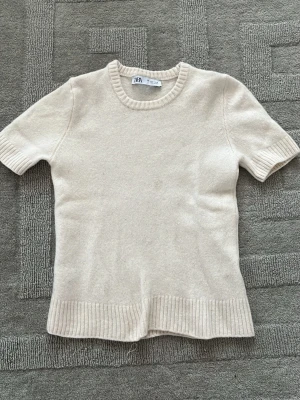 Vit kortärmad stickad ulltröja - Supermjuk vit stickad ulltröja från Zara med korta ärmar och ribbade detaljer vid hals, ärmslut och nederkant. Perfekt till jeans eller kjol för en clean och enkel look. Storlek S men är liten i storleken så typ XS.