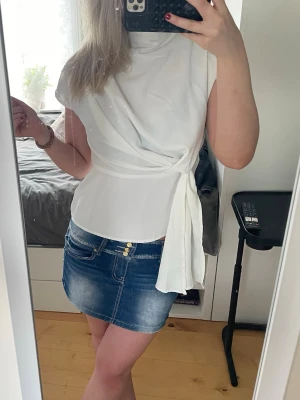Vit omlottblus med knyt i midjan - Snygg vit omlottblus med knytband i midjan och hög hals. Blusen har korta ärmar och en stilren, feminin passform som framhäver midjan. Perfekt att matcha med jeans eller kjol 🥰