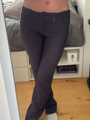 Lågmidjade kostymbyxor - Snygga gråa kostymbyxor med raka ben och dekorativa knappar i midjan. Byxorna har fickor bak och en stilren design som passar perfekt till en trendig look. Materialet känns mjukt och följsamt, vilket gör dem bekväma att bära hela dagen.