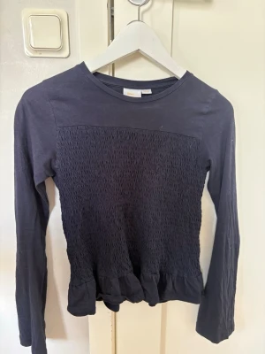 Mörkblå långärmad topp med rynk - Mörkblå långärmad topp med rynkdetaljer över bröst och rygg samt volang längst ner. Rund halsringning och mjukt material som känns skönt mot huden. Perfekt för en chill och stilren look.