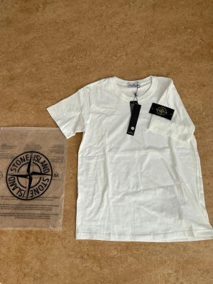 Vit Stone Island t-shirt i bomull - Helt ny! Vit t-shirt från Stone Island i mjuk bomull med klassisk rund hals och korta ärmar. På vänster ärm sitter den ikoniska Stone Island-loggan. Storlek SMALL och stämmer bra i storleken. Kan skicka bilder hur den sitter på i chatten och vikt och längd 