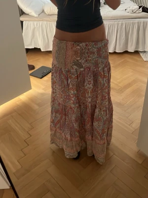 Mönstrad långkjol i boho-stil - Säljer en färgglad långkjol med bohemiskt mönster i rosa, rött och beige toner. Kjolen har en smockad midja och faller snyggt i flera lager. Perfekt för dig som gillar en avslappnad och trendig look.