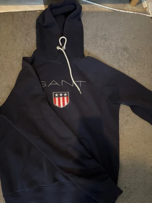 Mörkblå hoodie från GANT - Snygg mörkblå hoodie från GANT med vit dragsko i huvan och broderad logga samt sköld med stjärnor och ränder på bröstet. Klassisk känguruficka framtill och mjukt material som känns skönt mot huden. Perfekt för en avslappnad och sportig stil.