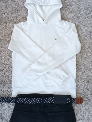 Vit hoodie från Gant med broderad logga - Snygg vit hoodie från Gant med liten broderad logga på bröstet. Klassisk modell med huva och känguruficka framtill. Mjuk bomullskänsla och ribbade muddar vid ärmslut och nederkant. Perfekt för en clean och stilren look.