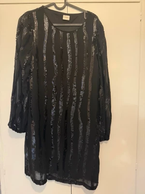 Svart paljettklänning från Jacqueline de Yong - Svart kort klänning från Jacqueline de Yong med långa ärmar och glittriga paljettränder. Klänningen har en rund halsringning och är tillverkad i polyester, perfekt för dig som vill sticka ut med en snygg och trendig look.