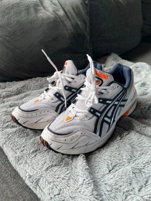 Asics vita sneakers med marinblå detaljer - Säljer ett par fräscha Asics sneakers i vitt med marinblå och orange detaljer. Skorna har klassisk mesh- och syntetöverdel, tydliga Asics-loggor på sidorna och en chunky sula med orange inslag. Perfekta för en sportig och trendig look.Är väldigt bra spring och promenadskor och är väldigt bra för kroppen då den har gel sulor.Är ändrat använda en gång så är som gott som nya. Lite svarta hår i sulan kan förekomma då hemmet har en hund, men dem är tvättade i tvättmaskin och det är inget som känns. 
