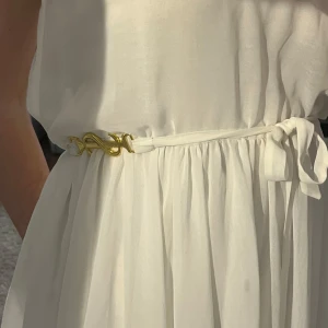 Vit kortklänning från Ida Sjöstedt - Superfin vit kortklänning från Ida Sjöstedt med guldiga detaljer på axelbandet och ett knytband i midjan. Klänningen har en luftig och flowy passform, perfekt för varma dagar. Stilren och elegant look med tunna axelband. Står inte storlek någonstans men gissar på S