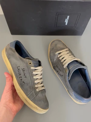Saint Laurent sneakers - Gråa låga sneakers från Saint Laurent med vit sula och vita skosnören. Skorna har handskriven Saint Laurent-logga på sidan och blå detaljer vid hälen. Tillverkade i mocka och har en klassisk, stilren siluett.