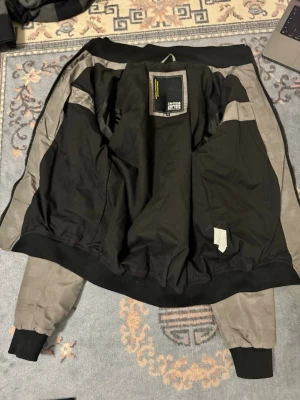 Grå bomberjacka från Jack & Jones - Snygg grå bomberjacka från Jack & Jones med svarta muddar och detaljer. Jackan har dragkedja, två fickor med knappar och en ärmficka med dragkedja. Perfekt för dig som vill ha en stilren och trendig look.