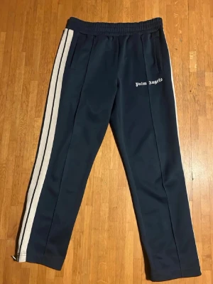 Palm Angels blå byxor - Palm Angels byxor i blå med vita sidoränder och logga på vänster ben. Byxorna har resår i midjan och raka ben. Tillverkade i ett mjukt och följsamt material som ger en sportig och trendig look.