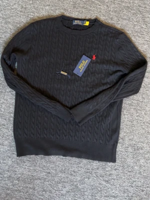 Svart kabelstickad tröja Polo Ralph Lauren - Kabelstickad svart tröja från Polo Ralph Lauren med rund halsringning och röd broderad logga på bröstet. Tröjan har långa ärmar och ribbade muddar vid ärmslut och nederkant. Perfekt för en stilren och klassisk look.