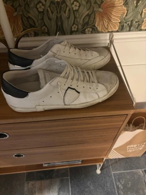 Vita Philippe Model sneakers med svart detalj - Snygga vita sneakers från Philippe Model med svart häl och klassisk sköld på sidan. Använt skorna fåtal gånger så de är i väldigt bra skick. Perfekta för en stilren och trendig look.