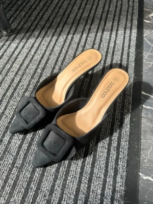 Svarta pumps med rosett från Boohoo - Snygga svarta pumps från Boohoo med spetsig tå och dekorativ rosett framtill. Skorna har öppen häl och smal klack, vilket ger en elegant siluett. Ovandelen är i textil och innersulan är beige för en snygg kontrast.