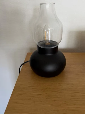 Svart bordslampa med glaskupa - Säljer en stilren bordslampa med rund svart fot i metall och en genomskinlig glaskupa. Lampan har en klassisk form som påminner om en oljelampa och passar perfekt som mysbelysning. Strömkabeln är svart och lampan har en synlig glödlampa.