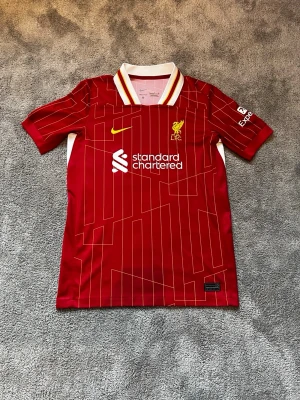 Liverpool hemma matchtröja 2024/25  - Säljer en röd Liverpool FC matchtröja från Nike med vita och gula detaljer. Tröjan har klubbmärke och sponsorlogga broderat på bröstet, samt mönstrade linjer över hela plagget. Klassisk krage med ränder och korta ärmar. Perfekt för fotbollsfans!