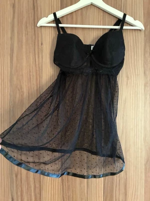 Svart spets babydoll med meshkjol - Snygg svart babydoll med vadderade kupor i spets och justerbara axelband. Kjolen är gjord i transparent mesh med små prickar och satinkant nedtill. Perfekt för dig som gillar romantiska och feminina detaljer.