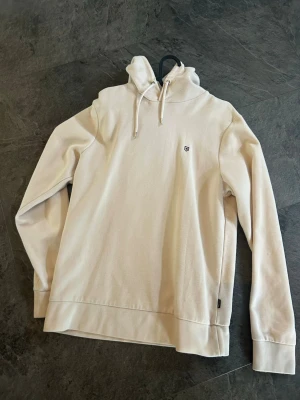 Beige hoodie från jack and jones - Snygg beige hoodie från jack and jones med liten broderad logga på bröstet. Klassisk modell med huva och dragsko, samt ribbade muddar vid ärmslut och nederkant. Perfekt för en avslappnad och stilren look.