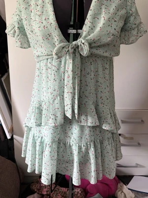 Mintgrön blommig kortklänning med volanger - Söt mintgrön kortklänning med små blommor, volanger och knyt framtill. Klänningen har korta ärmar med volangdetaljer och omlottkänsla vid bysten. Perfekt för sommarens alla tillfällen.