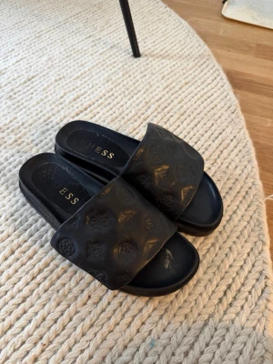 Svarta sandaler från GUESS - Svarta sandaler från GUESS med bred rem i präglat konstläder och diskret logomönster. Snygg chunky sula med räfflad undersida för extra grepp. Perfekta slides för en trendig och avslappnad look.