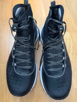 Svarta högskaftade sneakers från Under Armour  - Snygga svarta sneakers från Under Armour med högt skaft och vita sulor. Skorna har svarta och vita mönstrade skosnören, ventilerande meshpartier och detaljer i syntetmaterial. Perfekta för dig som gillar sportig och modern stil.