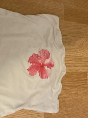 Vit tröja med en rosa blomma i mitten ingen arm - Vit t-shirt med en stor rosa blomma tryckt framtill. Enkel och fräsch design som passar perfekt till jeans eller shorts. Tillverkad i mjuk bomull för en bekväm känsla hela dagen.