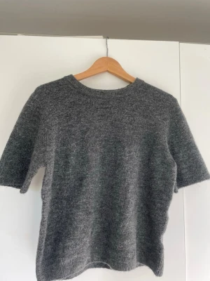 Grå stickad t-shirt i ullmix - Mysig grå stickad t-shirt med korta ärmar och rund hals. Tröjan har en enkel och stilren design med ribbstickade kanter vid ärmslut och nederkant. Perfekt för lager-på-lager och passar till många olika stilar.
