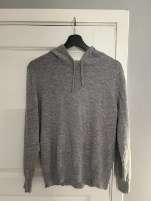 Grå stickad cashmere hoodie  - Mysig grå hoodie i mjukt stickat material med huva och dragsnören. Långärmad modell med ribbade muddar vid ärmslut och nederkant. Perfekt för en avslappnad och chill stil.