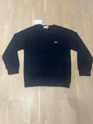 Mörkblå sweatshirt från Lacoste - Mörkblå sweatshirt från Lacoste med klassisk rund halsringning och det ikoniska gröna krokodilmärket broderat på bröstet. Tröjan har ribbade muddar vid ärmslut och nederkant. Perfekt för en stilren och avslappnad look.