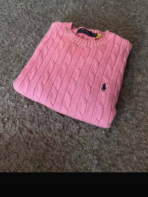 Rosa stickad tröja Polo Ralph Lauren - Snygg rosa kabelstickad tröja från Polo Ralph Lauren med rund halsringning och broderad logga på bröstet. Tröjan har ribbade muddar vid ärmslut och nederkant. Perfekt för lager-på-lager och en klassisk look.