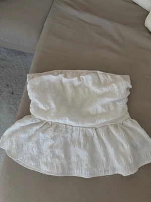 Vit axelbandslös bandeau topp - Söt vit bandeau topp med volangkant nedtill. Toppen är axelbandslös och har en lätt rynkad struktur i tyget som ger en avslappnad och somrig känsla. Perfekt att matcha med höga jeans eller kjol.