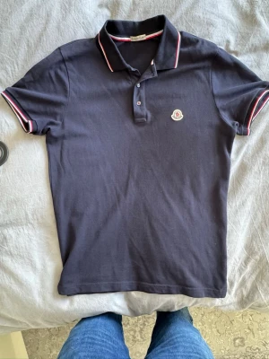 Moncler pikétröja mörkblå  - Snygg mörkblå pikétröja från Moncler med klassisk krage och knäppning framtill. Tröjan har röda och vita ränder på krage och ärmslut samt Moncler-logga på bröstet. Perfekt för en stilren och sportig look.